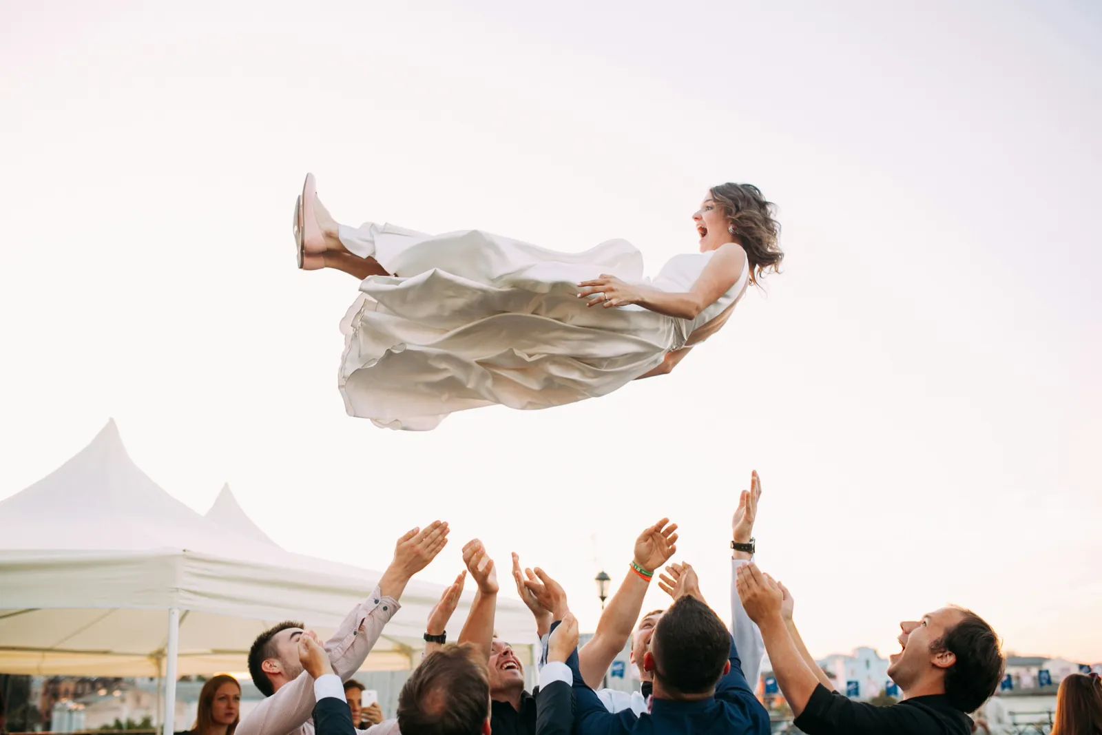 Guests lifting and tossing the bride in the air during an outdoor wedding celebration